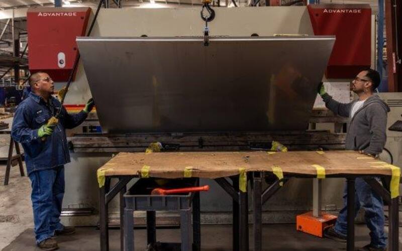 Press brake operators guiding formed steel panel with overhead crane